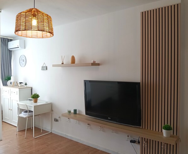 A modern living room with a flat-screen TV, a white desk, a chair, a hanging wicker lamp, and a wooden slat wall panel.