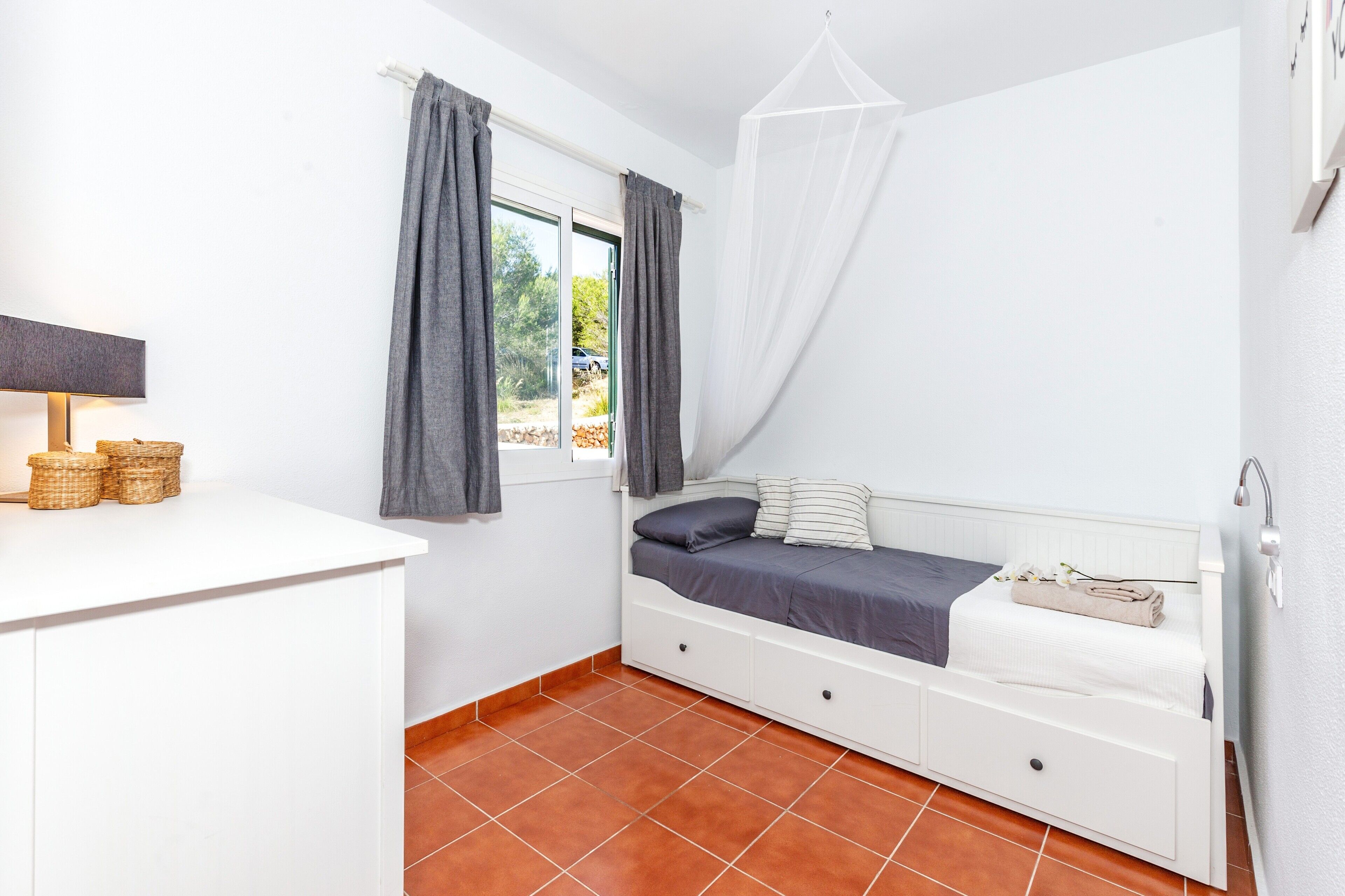 Een slaapkamer met een bed, een raam met gordijnen, een nachtkastje met manden, en een witte kast.