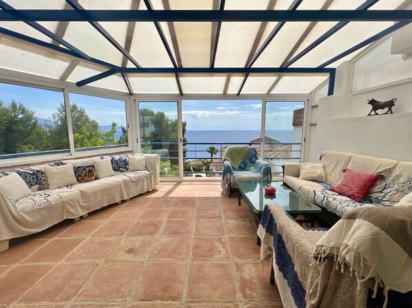 A sunlit room with a glass ceiling, two sofas, a glass table, and a view of the sea.