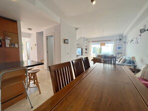 A spacious living area with a wooden dining table, chairs, a TV, and a balcony with a view.