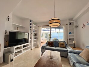 A living room with a TV, shelves, a sofa, and a view of the sea.
