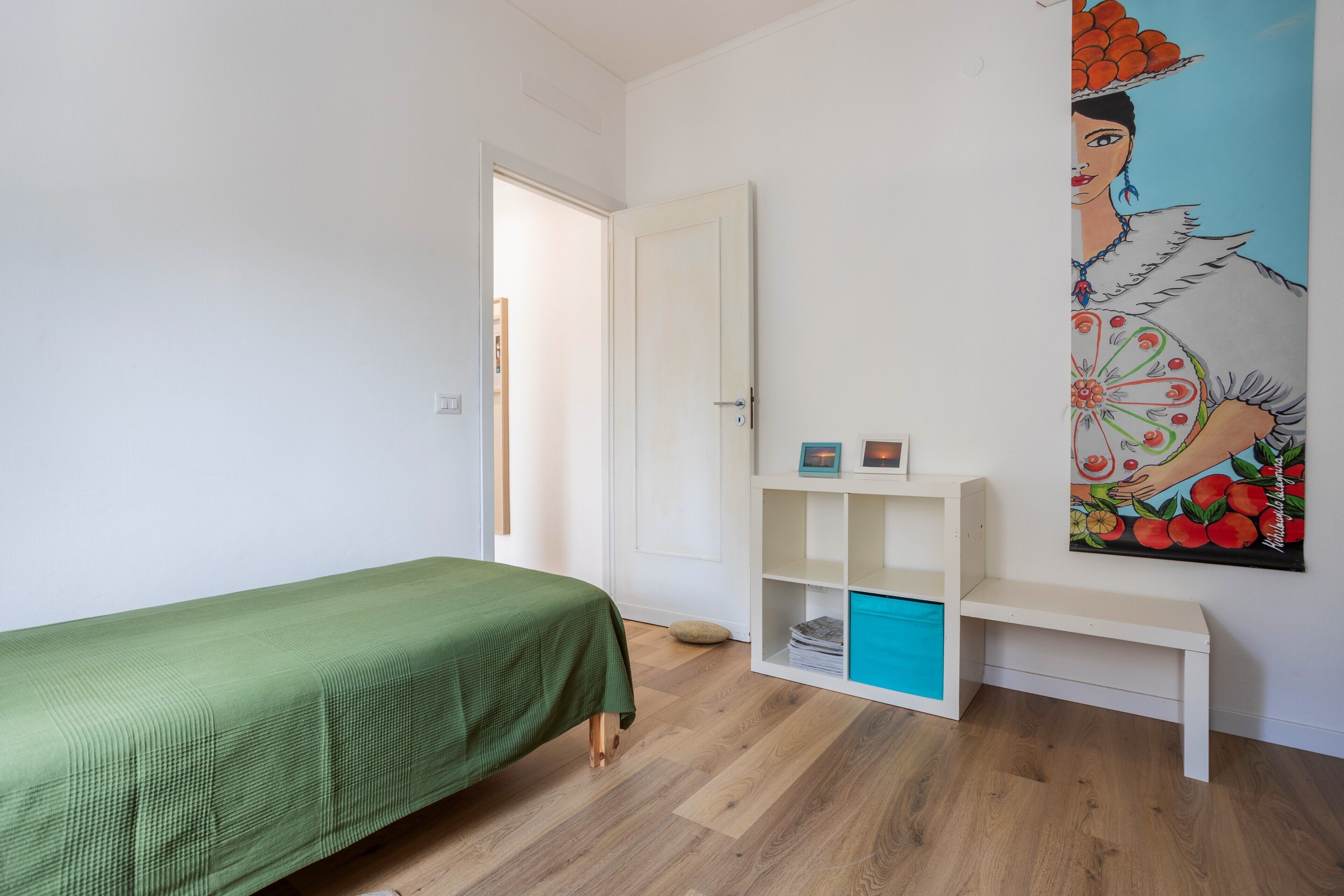 Una habitación con una cama verde, un armario blanco, un banco blanco y un cuadro colorido en la pared.