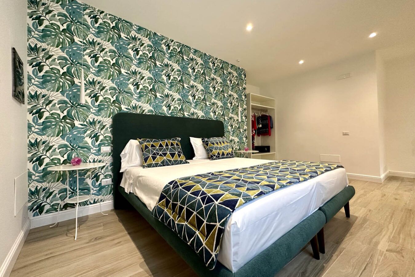 A bedroom with a large bed, a nightstand, a wardrobe, and a wall with a leafy pattern.