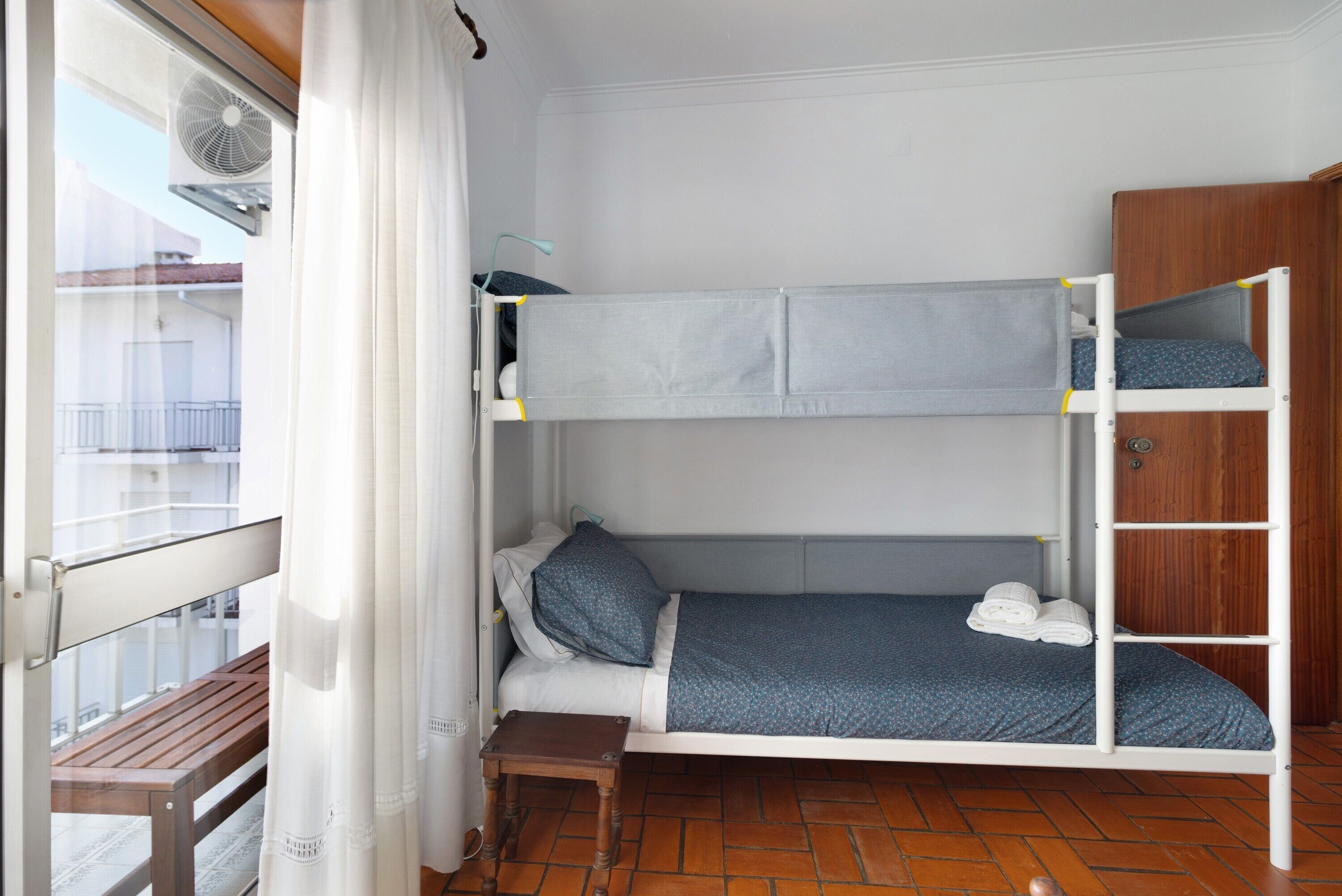 Een slaapkamer met een stapelbed, een raam, een ventilator en een houten bank.