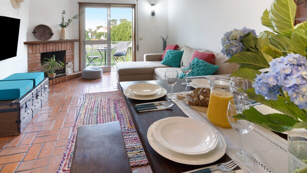 A dining area with a table set for four, a fireplace, a sofa with cushions, and a view through a glass door.