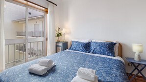 A bedroom with a bed, a nightstand, a lamp, and a view of a balcony.