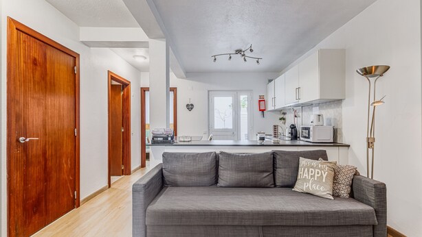 A modern living room with a grey sofa, wooden doors, and a kitchen in the background.
