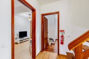 Um corredor com escadas de madeira, parede branca, extintor de incêndio e uma televisão ao fundo.