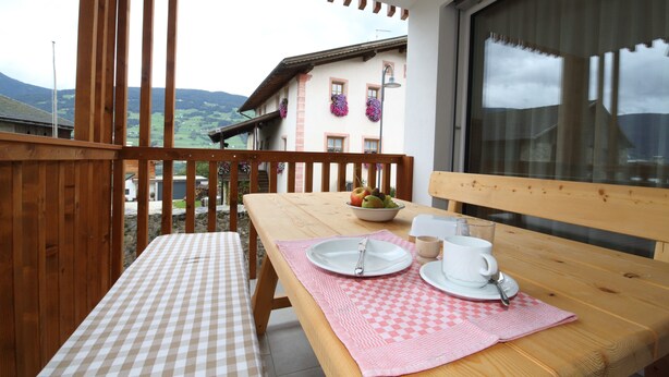 Un balcon en bois agrémenté d’une nappe à carreaux, d’un banc en bois et d’une table où sont disposés une assiette, une tasse et un bol de fruits.