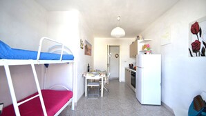 Una habitación compacta con literas, una kitchenette pequeña, un comedor y un refrigerador blanco.