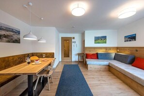 Un intérieur moderne avec une table à manger en bois, un canapé avec des coussins, des luminaires suspendus et une vue sur l’extérieur.