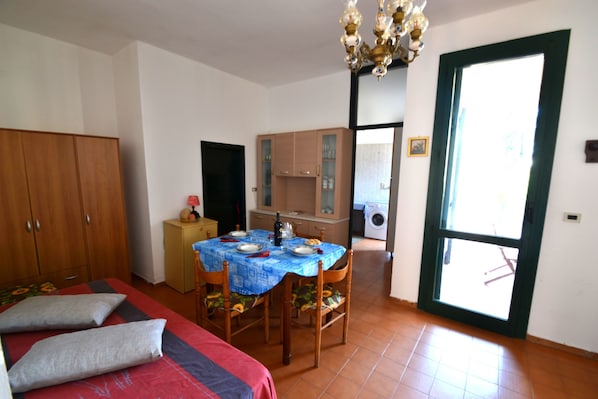A bedroom with a bed, a dining table with chairs, a kitchen area, and a large window.