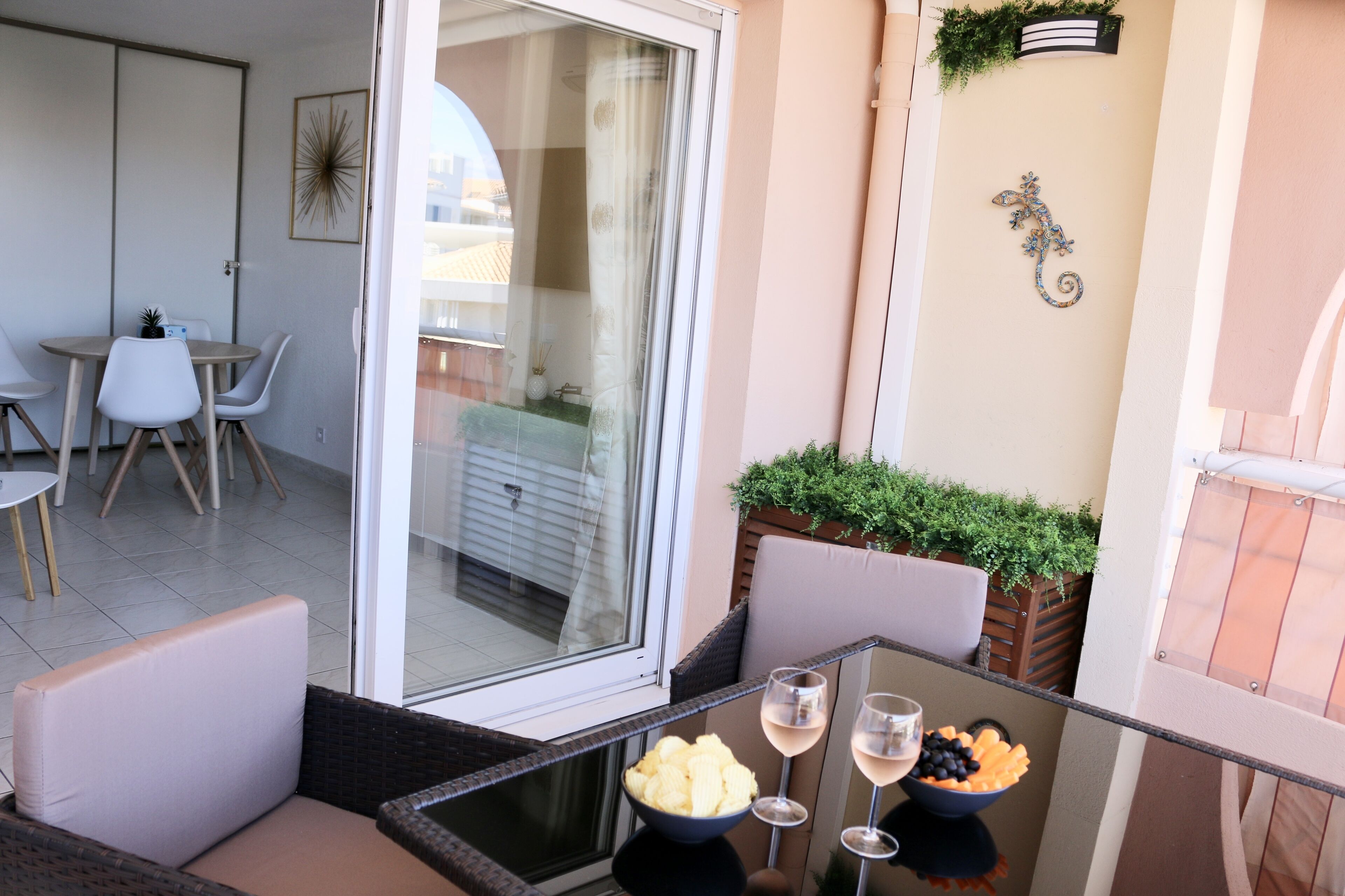 A balcony with wicker furniture, a glass table with a bowl of fruit and two glasses of wine, and a view of the cityscape through a glass door.