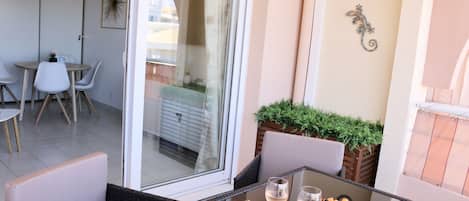 A balcony with wicker furniture, a glass table with a bowl of fruit and two glasses of wine, and a view of the cityscape through a glass door.
