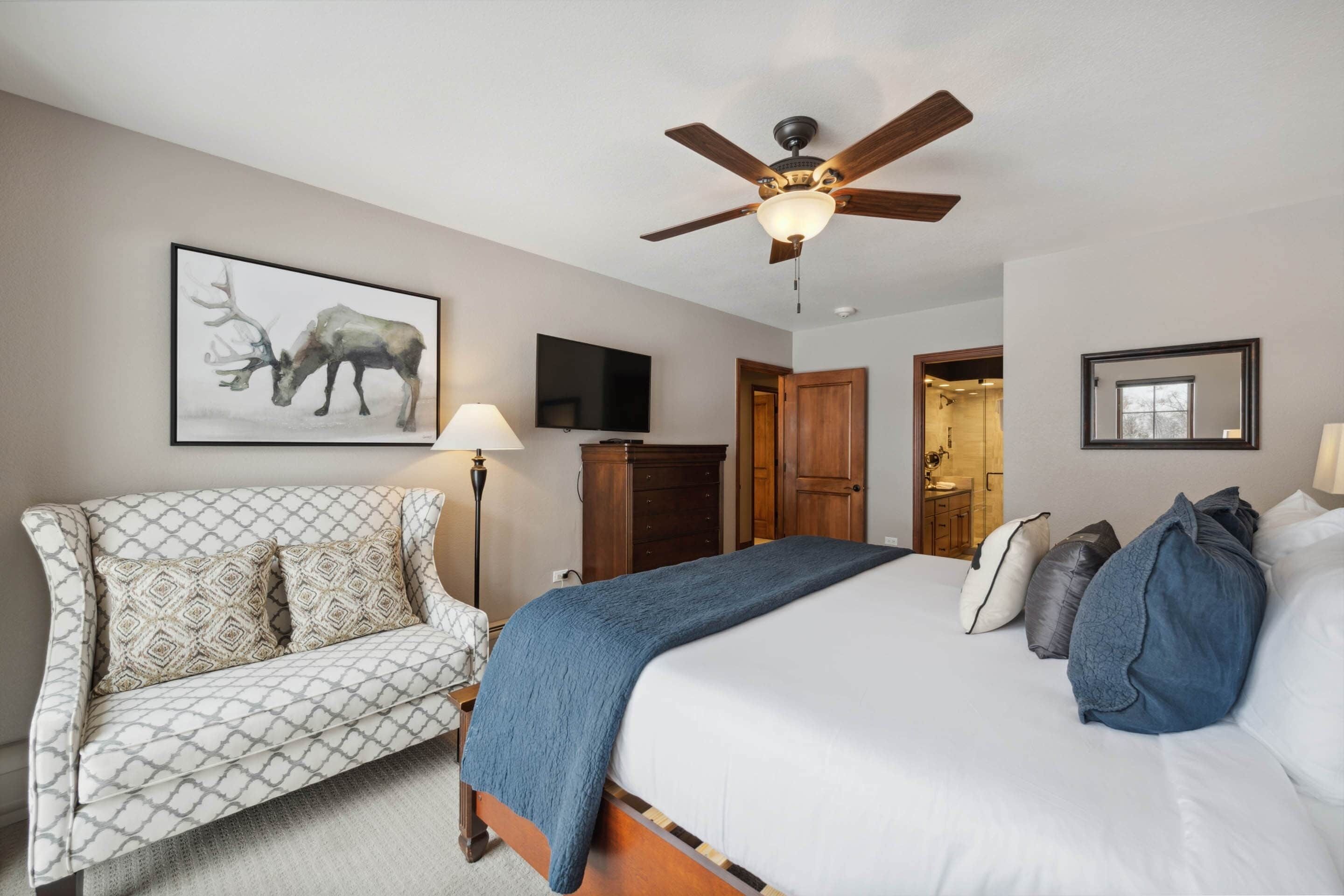 A bedroom with a bed, a sofa, a ceiling fan, a television, and a bathroom visible through an open door.