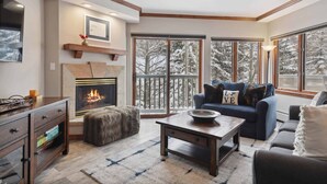 A cozy living room with a fireplace, a sofa, a coffee table, and large windows with a snowy view.
