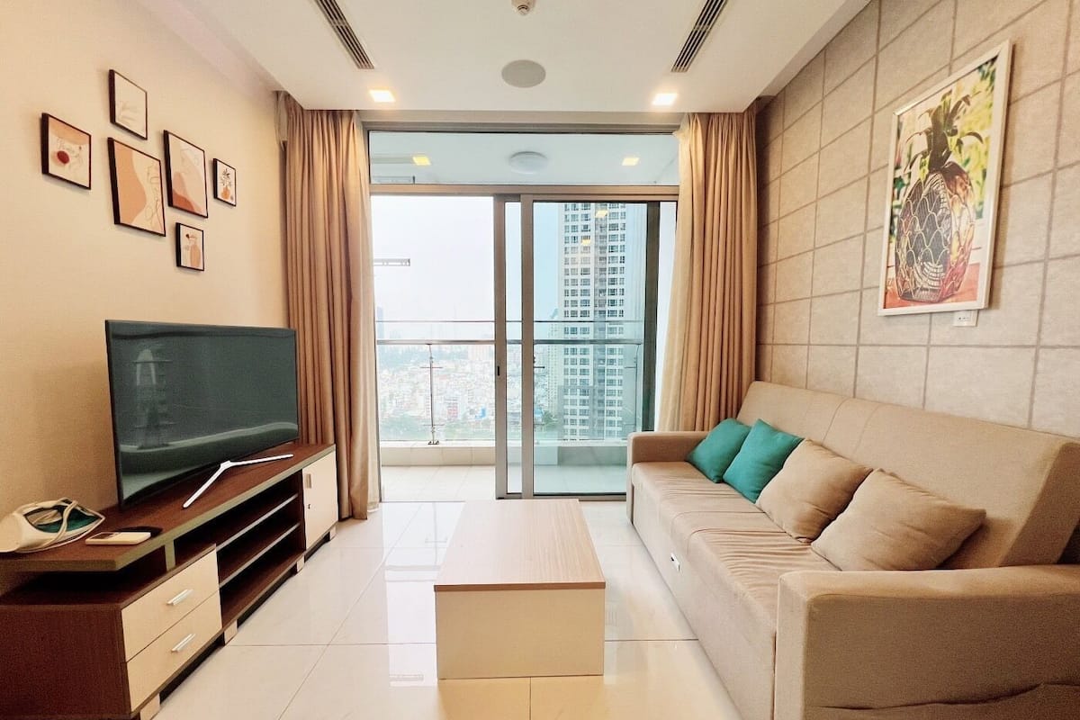 A modern living room with a flat-screen TV, a sofa with green cushions, a coffee table, and a city view through large windows.