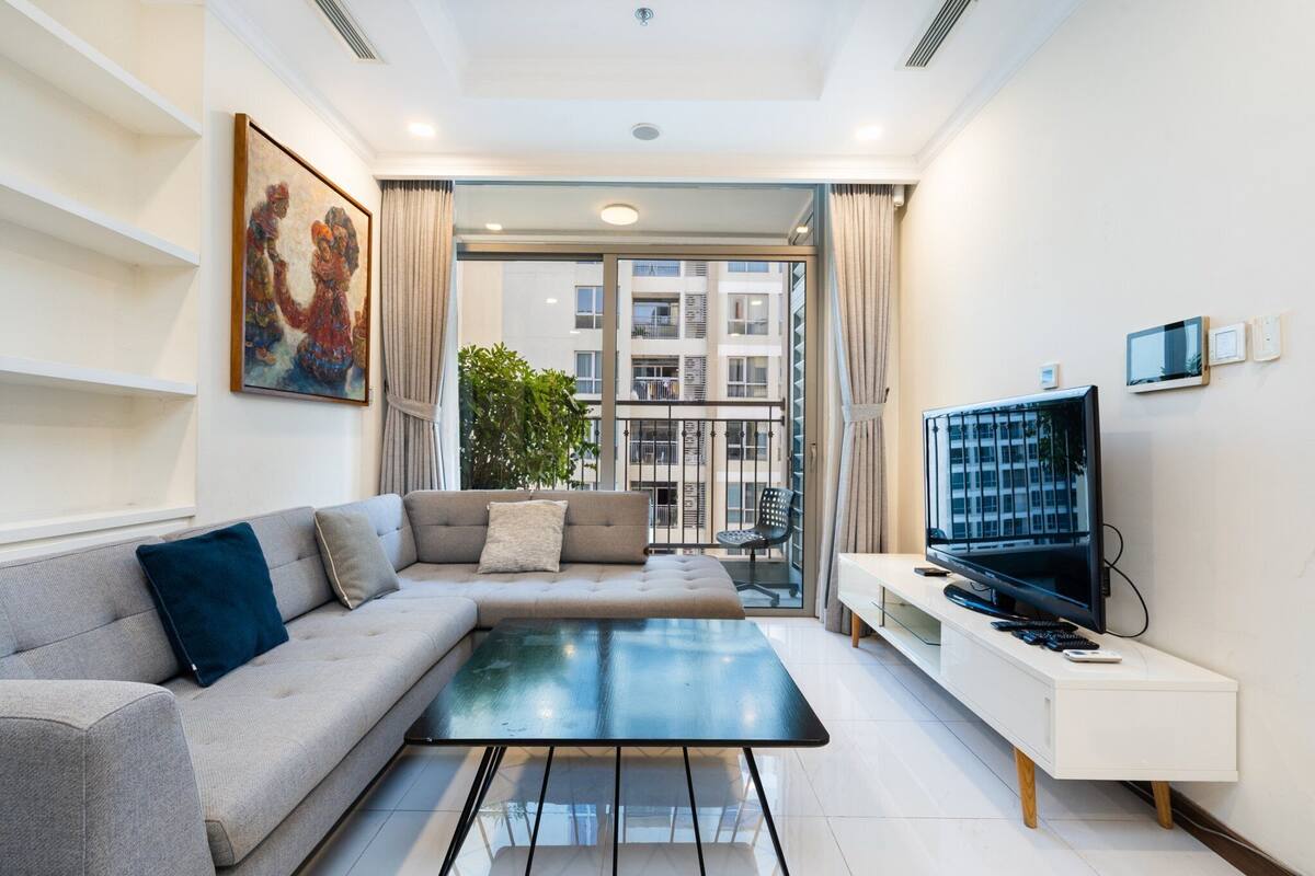 A modern living room with a grey sofa, a glass coffee table, a flat-screen TV, and a large window offering a view of a building.