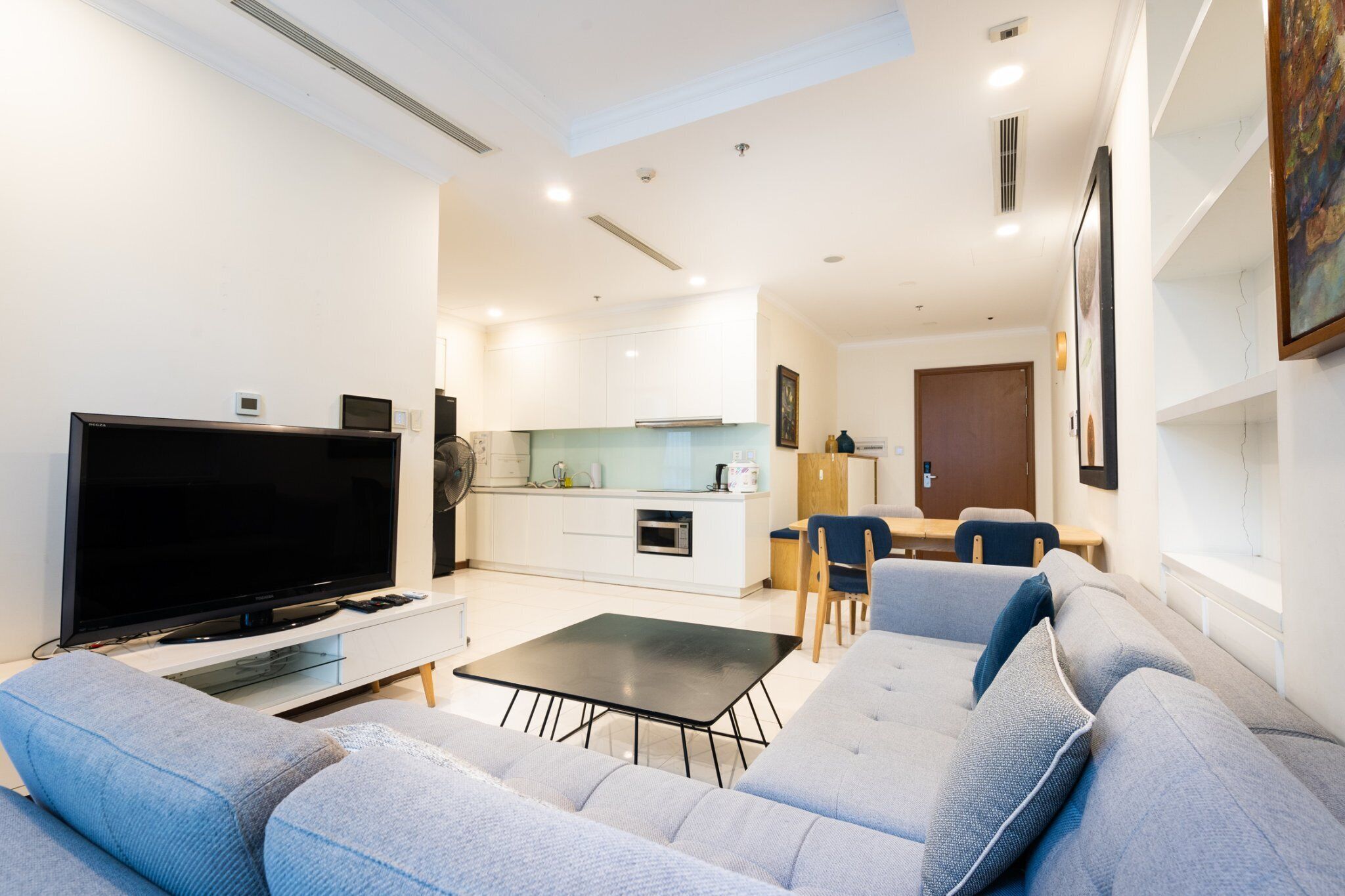 A modern living room with a flat-screen TV, grey sofa, and a small dining area with a table and chairs.