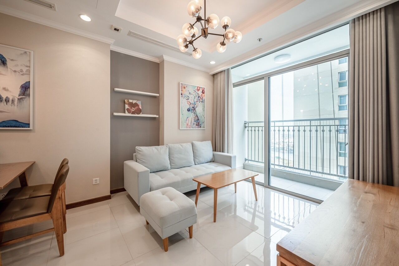 A modern living room with a sofa, ottoman, and coffee table. There is a dining table with chairs, a chandelier, and a large window with a view of buildings.