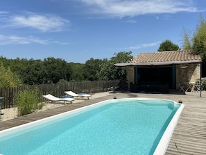 A rectangular swimming pool with a wooden deck, lounge chairs, and a stone building with a tiled roof.