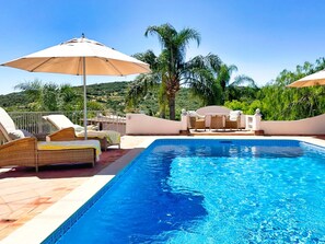 Una piscina con sillas de descanso y sombrillas, rodeada de palmeras y un paisaje montañoso.