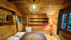 Een houten kamer met een bed, een hanglamp, een plank met spullen en een raam met gordijnen.