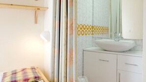 A bathroom with a white sink, a mirror, and a toilet. There is a small bed with a plaid blanket.