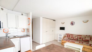 A compact kitchen with white cabinets, a sink, and a small refrigerator. There is a dining area with a brown table and a sofa with patterned cushions.
