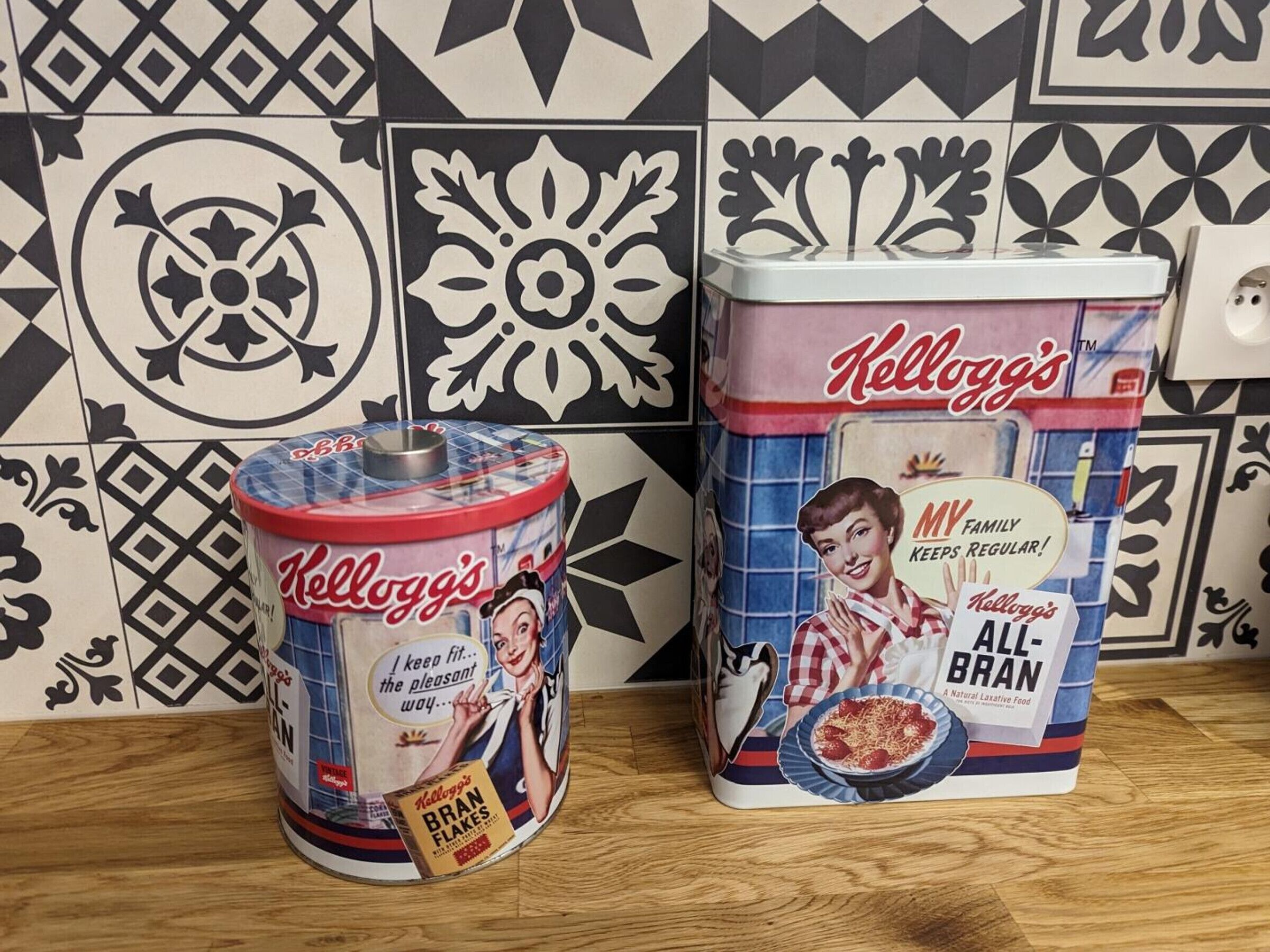 Two vintage Kellogg's All-Bran cereal containers on a wooden surface with a patterned tile wall in the background.