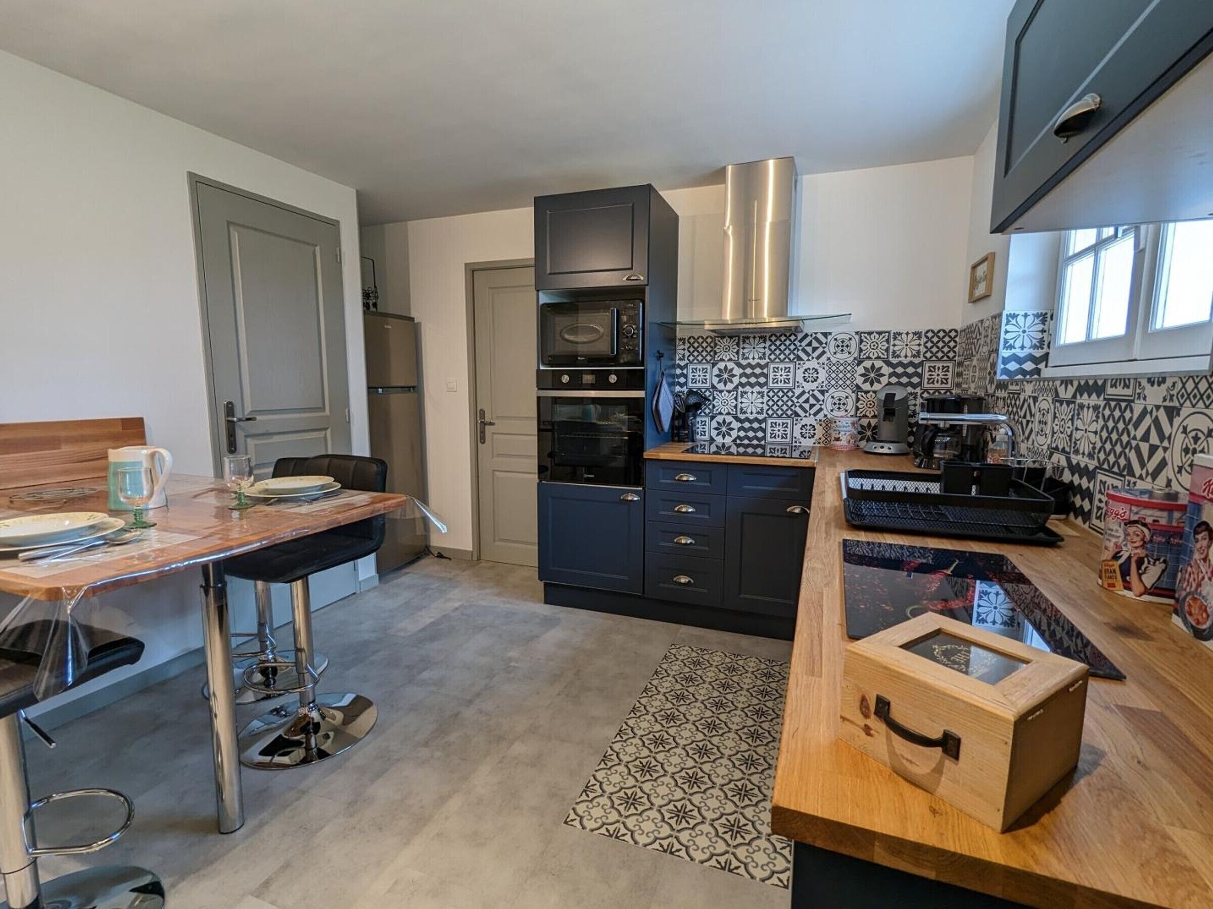 A modern kitchen with a wooden dining table, black cabinets, a stainless steel range hood, and patterned backsplash tiles.