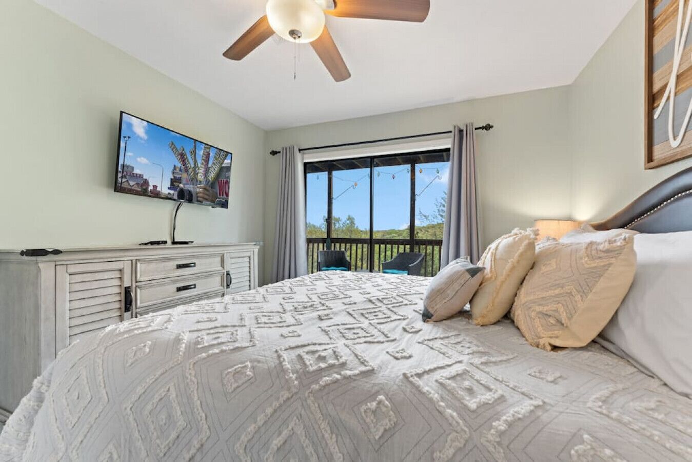 Un dormitorio con cama, ventilador de techo, televisor sobre una cómoda y balcón con vistas.