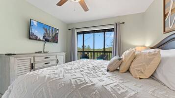 Un dormitorio con cama, ventilador de techo, televisor sobre una cómoda y balcón con vistas.