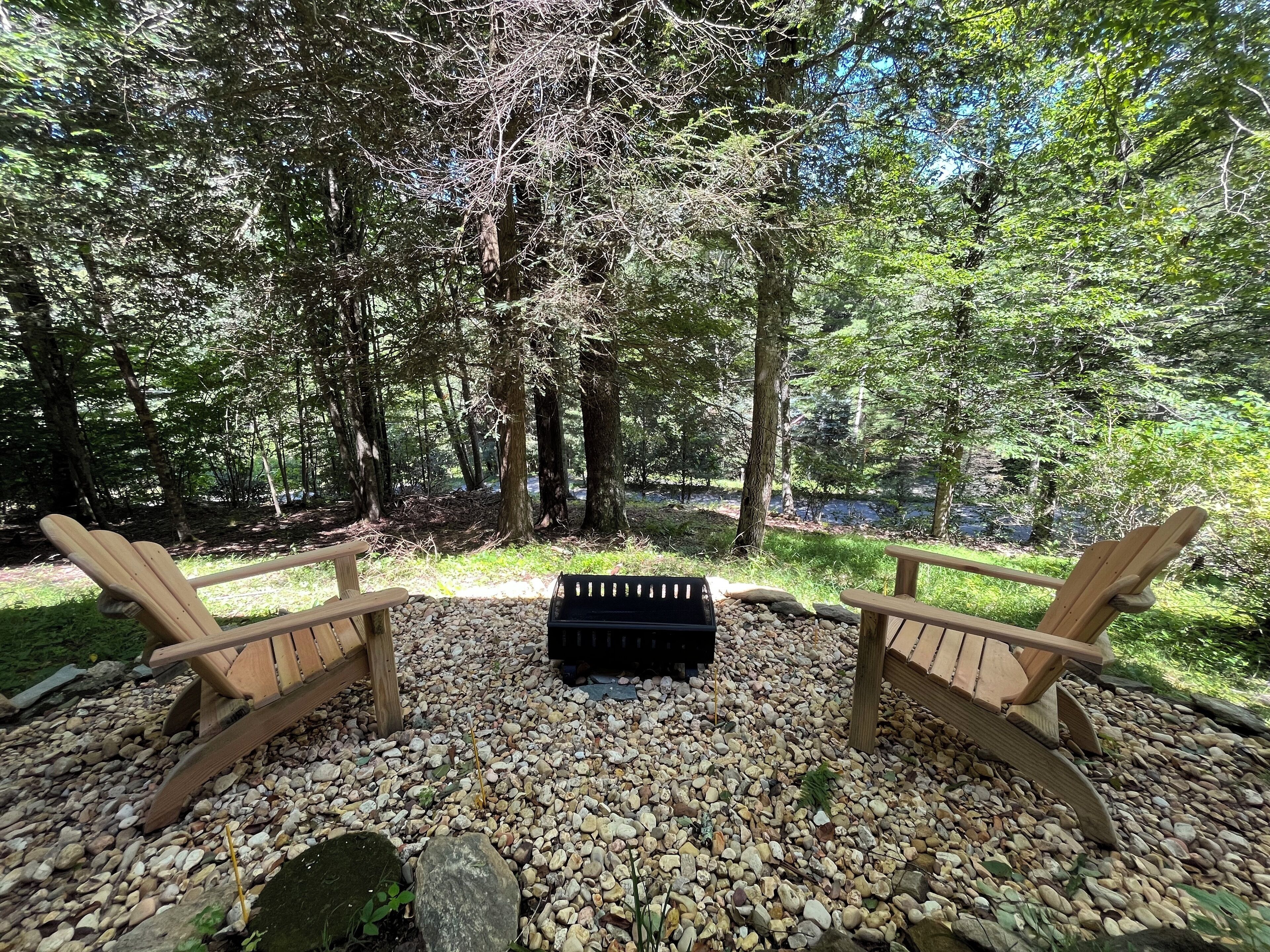Un espace aménagé avec deux fauteuils Adirondack en bois et un barbecue noir au centre, entouré d’un sol recouvert de petits graviers et d’arbres denses.