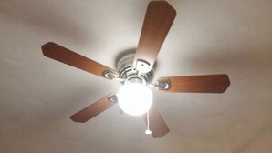 Un ventilador de techo con aspas de madera y una lámpara en una habitación con ventana, cortina y una lámpara de pared.