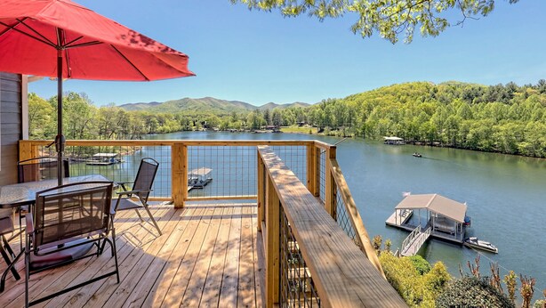 A wooden deck with a red umbrella, overlooking a lake with boats and a dock.
