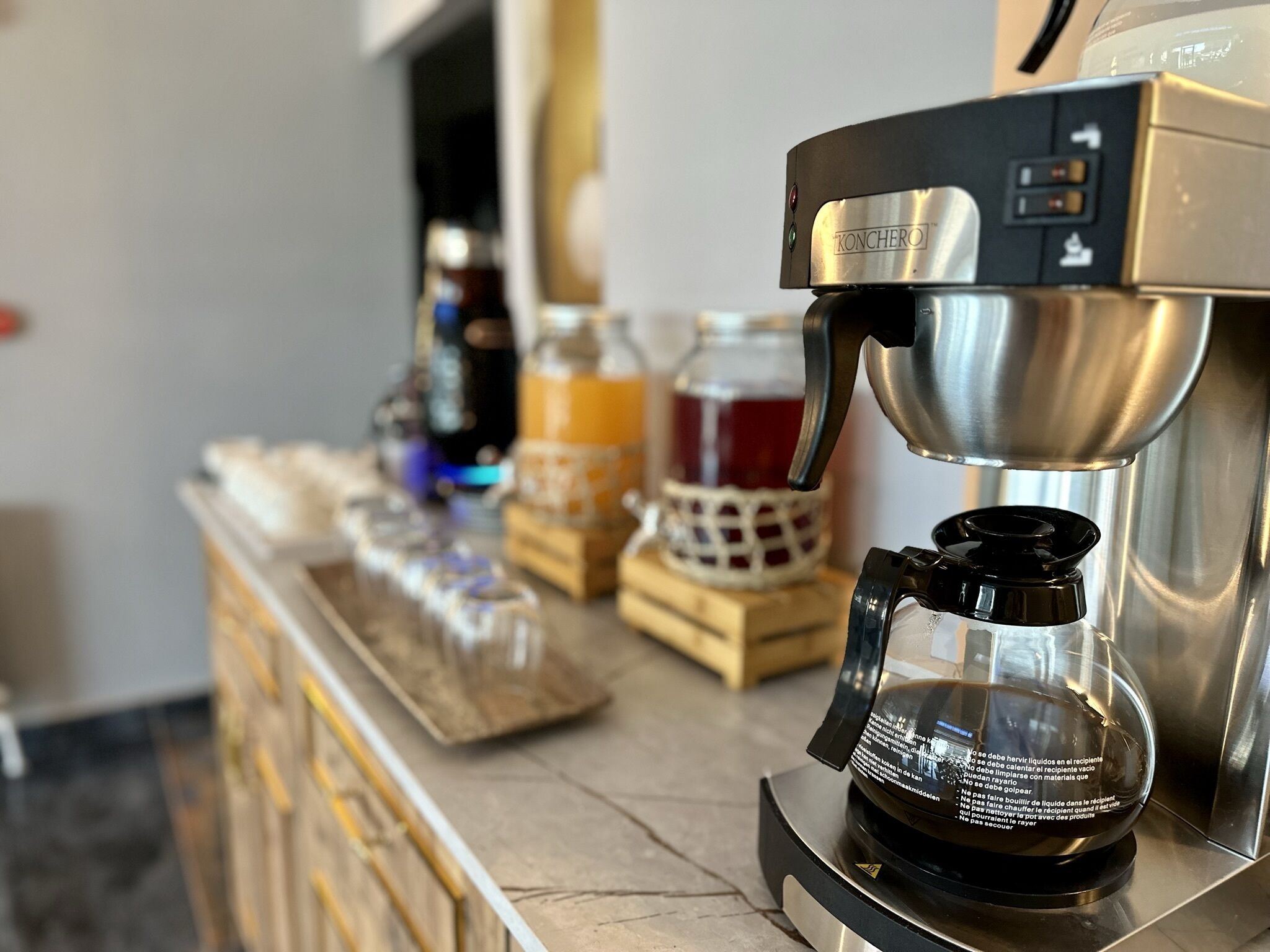 Una cafetera con jarra de vidrio sobre una encimera de mármol, un frasco de miel y una botella de miel al fondo.