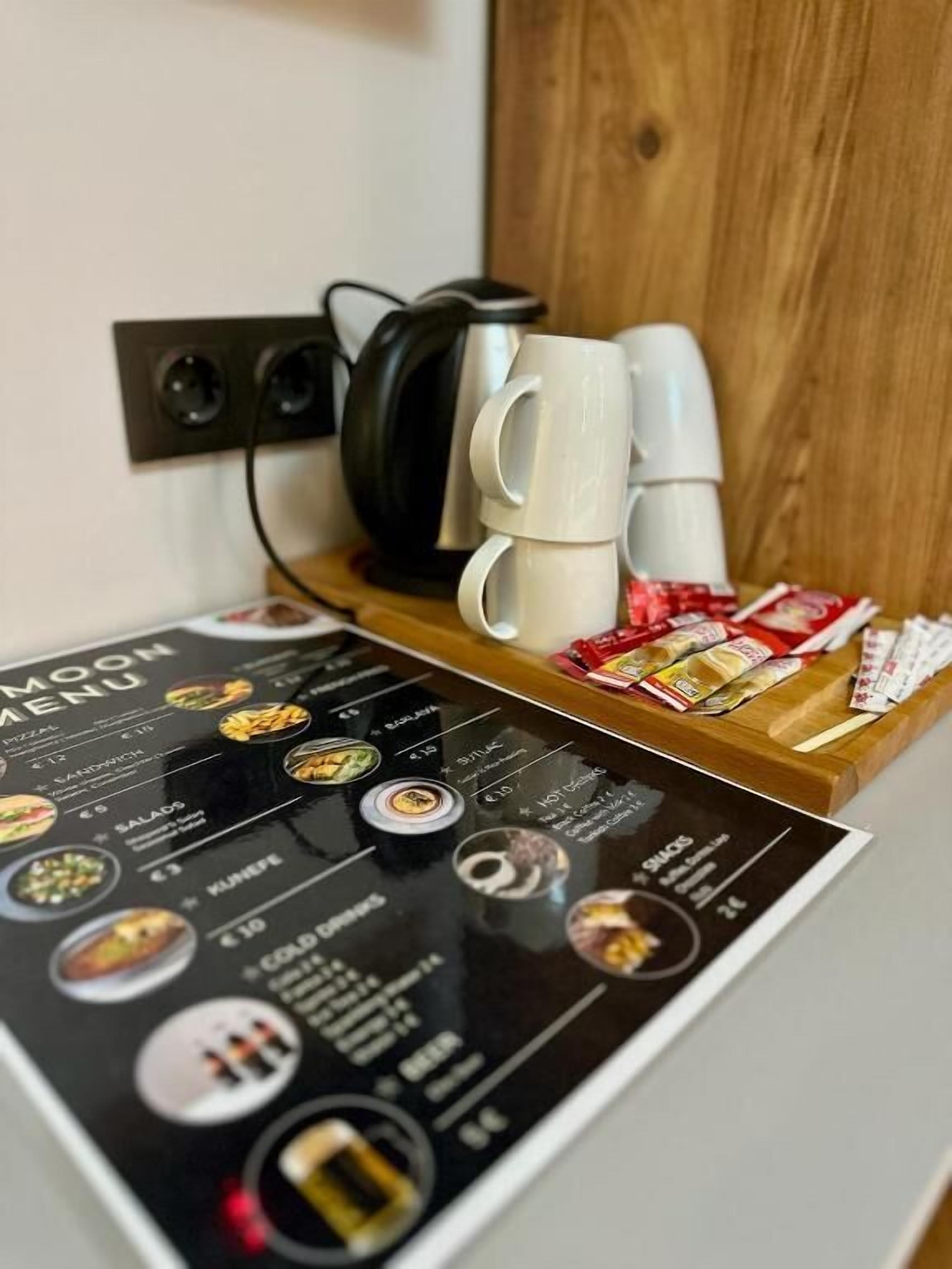 A menu board with food items and prices, a stack of white mugs, and a black kettle on a wooden shelf.
