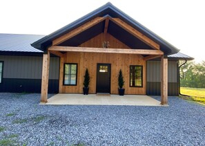 A wooden house with a covered porch, two potted evergreen trees, and a gravel driveway.