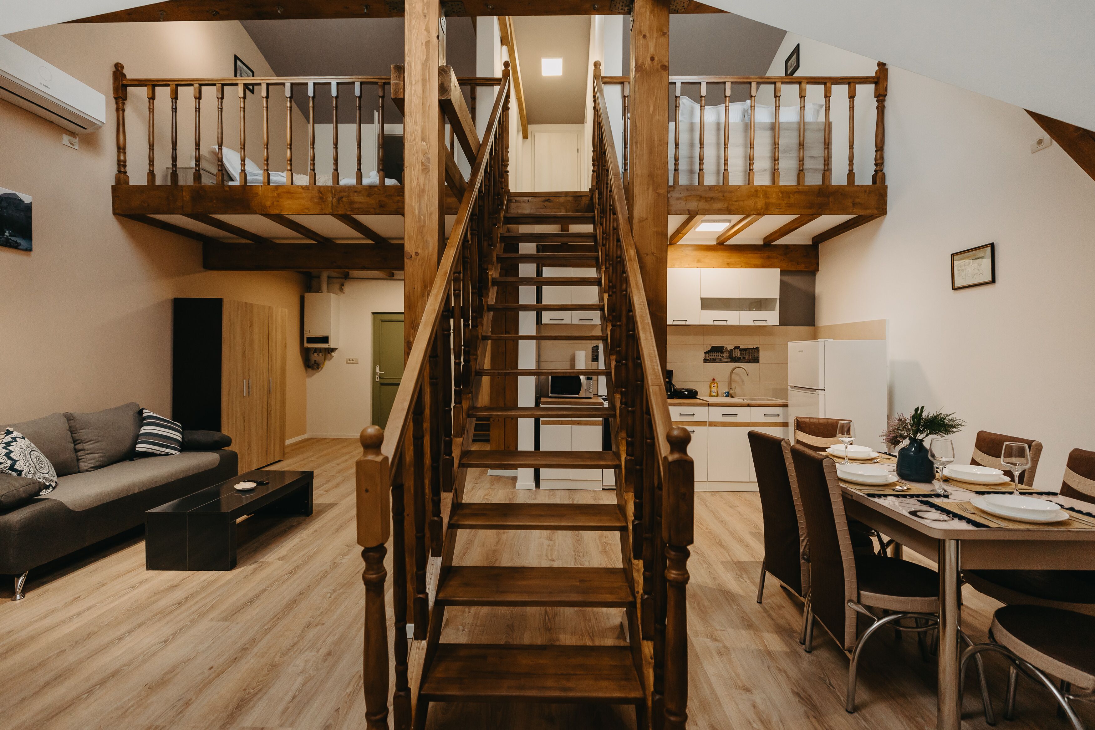 A modern loft apartment with a central wooden staircase connecting two levels. The living area features a dark sofa and a coffee table. The kitchen is compact with white cabinets and a dining table set for four.