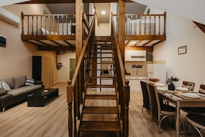 Un moderno apartamento tipo loft con una escalera de madera central que conecta dos niveles. La zona de estar presenta un sofá oscuro y una mesa de centro. La cocina es compacta, con armarios blancos y una mesa de comedor para cuatro personas.