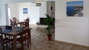 A dining area with a wooden table and chairs, a potted plant, and a painting of a coastal scene. - Appartement à Tiuccia, les Pieds Dans L'eau, Climatisé, Wifi (Tiuccia)