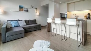 A modern living room with a grey sofa, a small dining area with white chairs, and a kitchen with a built-in oven and microwave.