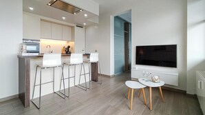 A modern kitchen with a built-in oven, microwave, and bar stools. There is a small dining table with two chairs and a TV mounted on the wall.