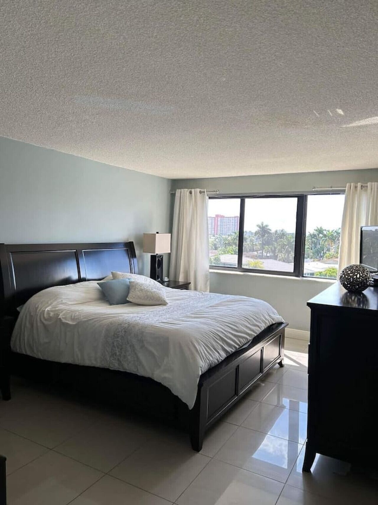 Un dormitorio con una cama grande, una mesita de noche, un aparador y una ventana con cortinas.
