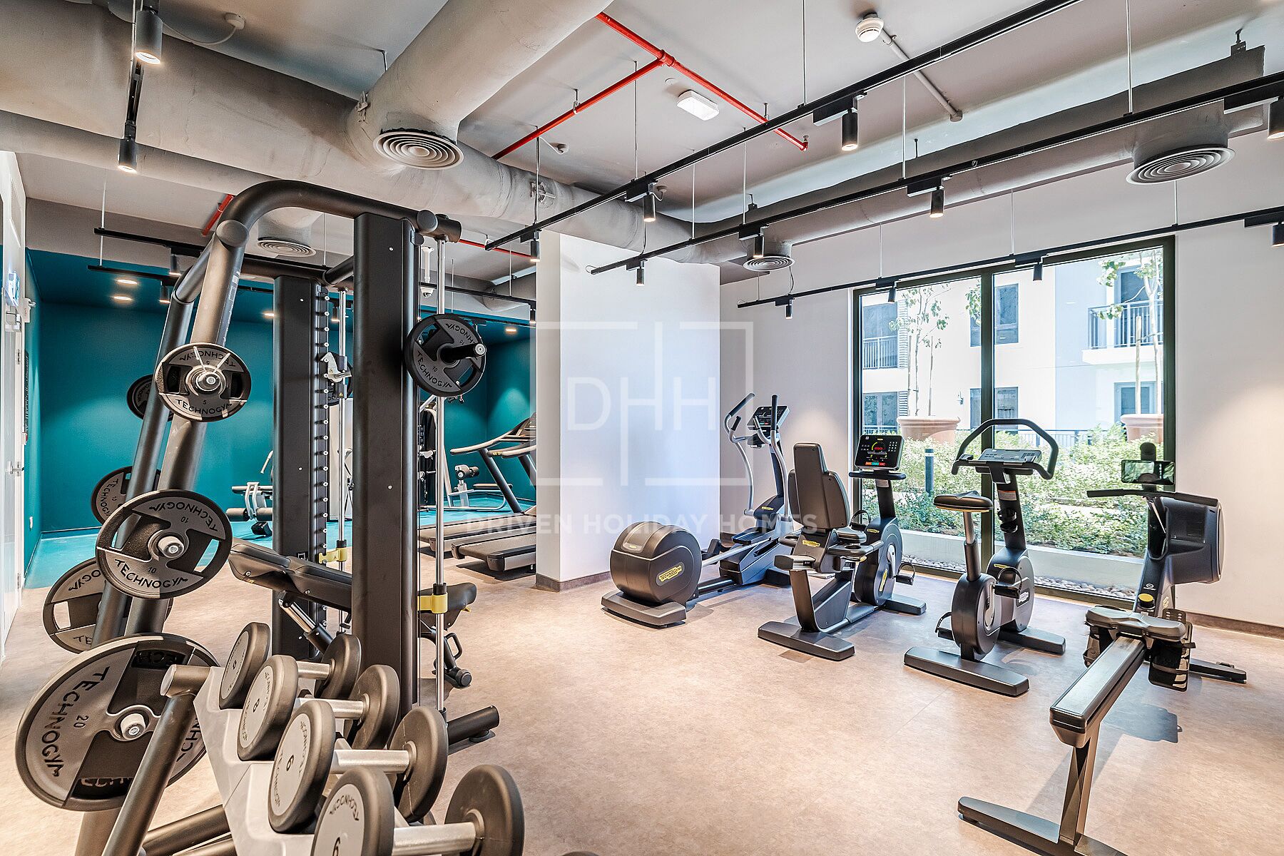 Une salle de sport équipée de divers appareils d’exercice, dotée de grandes fenêtres et d’un mur d’accent de couleur turquoise.