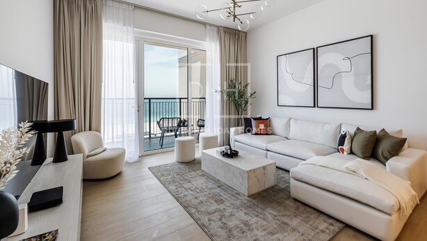 A modern living room with a white sofa, a coffee table, and a TV on a stand. There is a balcony with a view of the sky. - Full Sea View 1Bed Le Pont B1 in La Mer (Dubai)