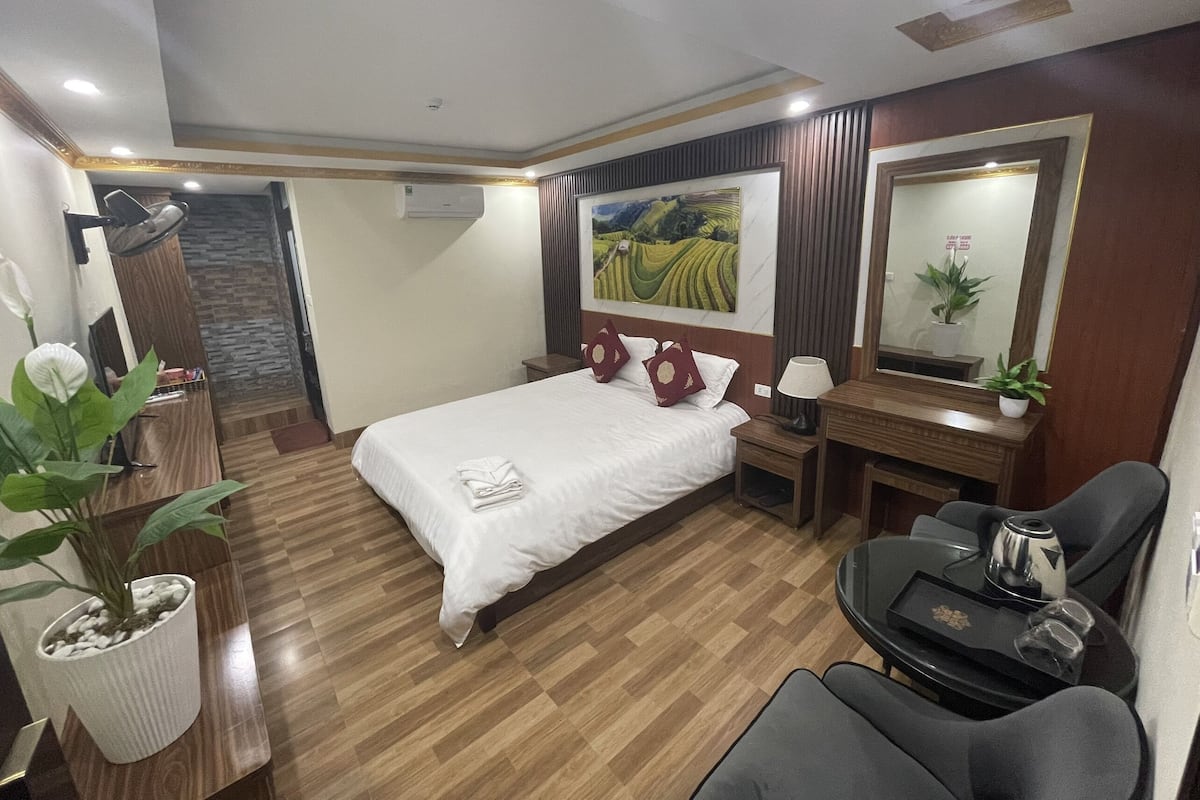 Een hotelkamer met een bed, een bureau met stoel, een kleine tafel met lamp, een spiegel en een potplant.