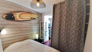 A bedroom with a wooden headboard, a bed with a white duvet, a wall-mounted surfboard decoration, and a patterned curtain. - Cozy 4-Person Apartment Steps from Beach, BBQ, WiFi & Parking (Capbreton)