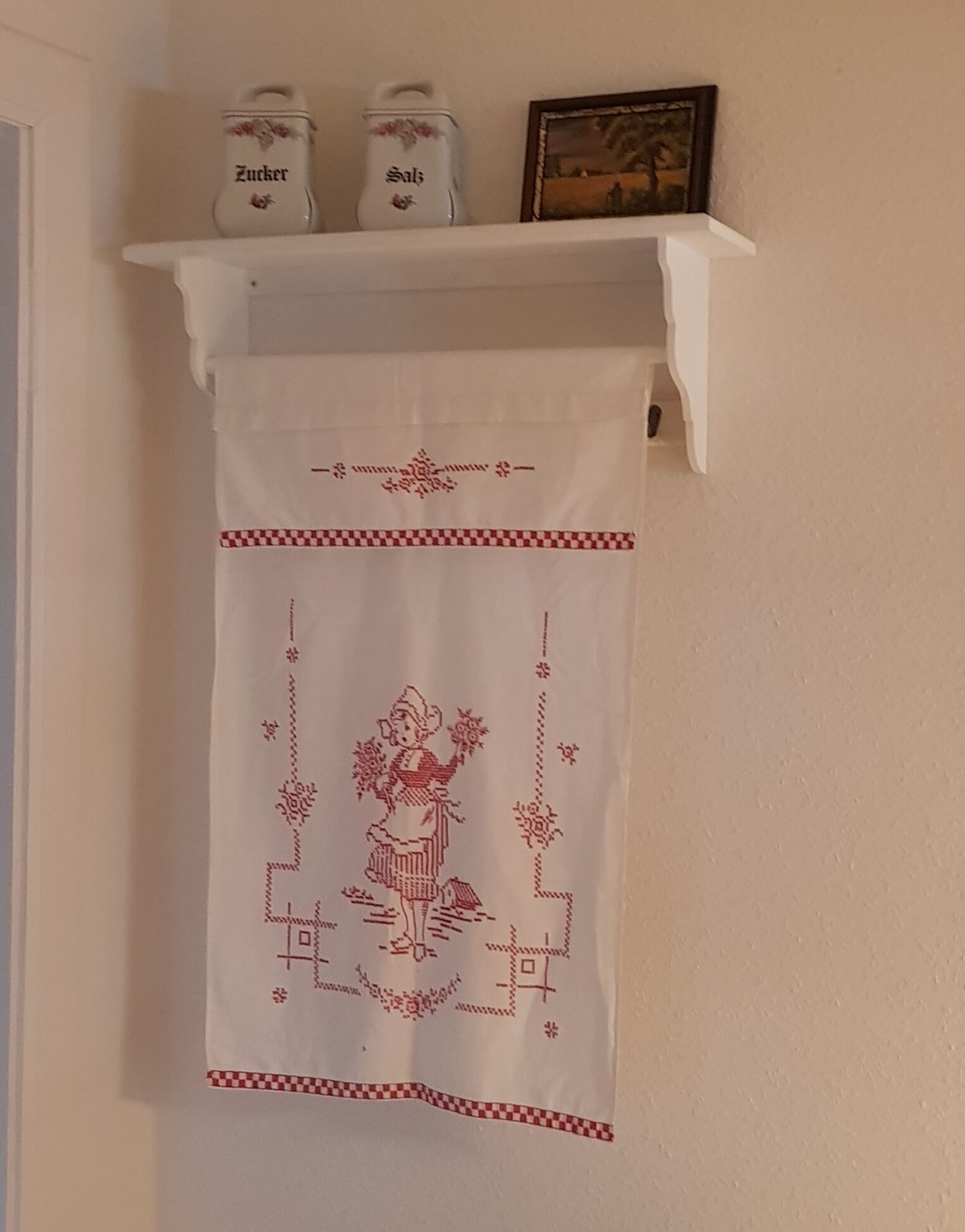 A kitchen area with a hanging white cloth featuring a red and white pattern. Above the cloth, there are two white containers labeled 'Zucker' and 'Salz' on a white shelf.